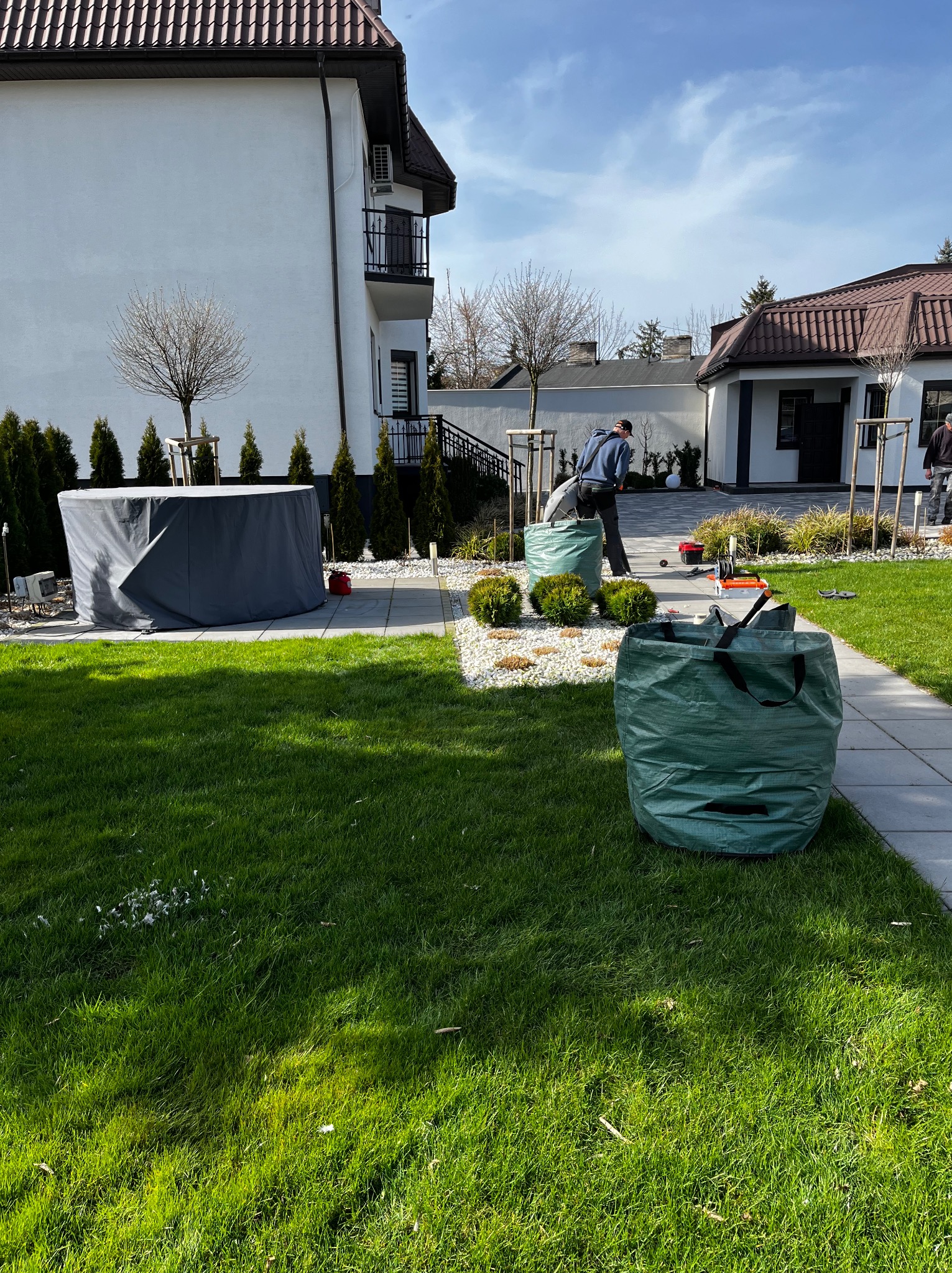 Prace ogrodnicze w zadbanym ogrodzie: pracownik w trakcie zbierania liści do worka, widoczne narzędzia ogrodnicze, trawnik, ozdobne krzewy i biały żwir.