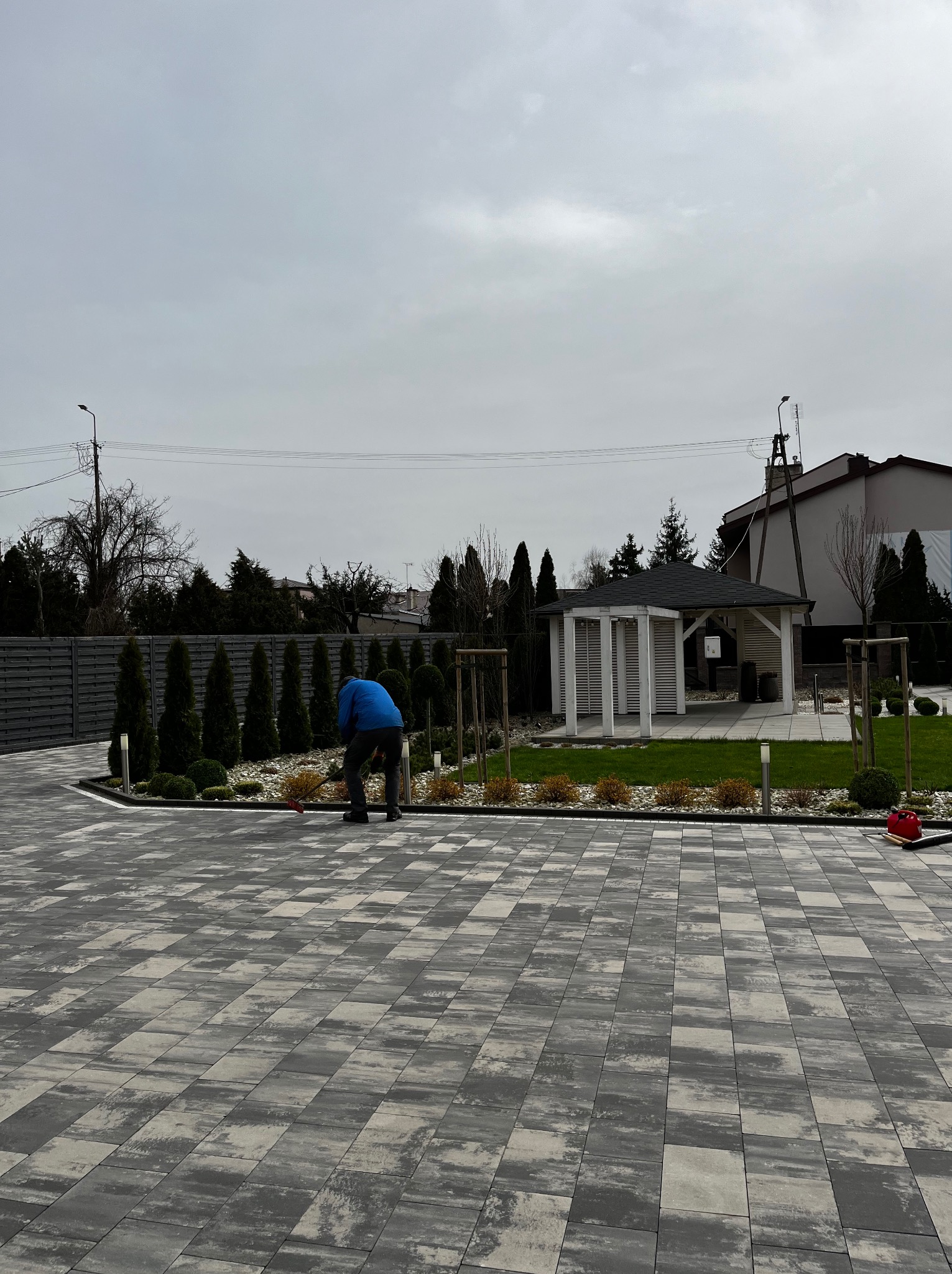 Mężczyzna w niebieskiej kurtce grabi liście na rabacie z kamyków, obok zadbanego trawnika i altany w ogrodzie, na pierwszym planie kostka brukowa.