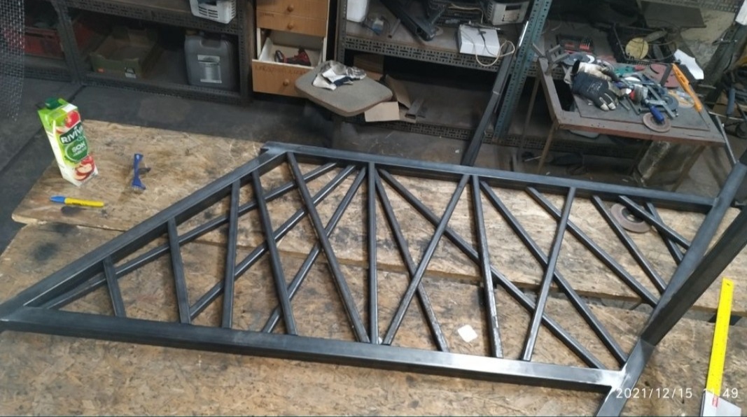 Metalowa konstrukcja schodów z geometrycznym wzorem na warsztatowym stole, widoczny karton soku, narzędzia i miara.