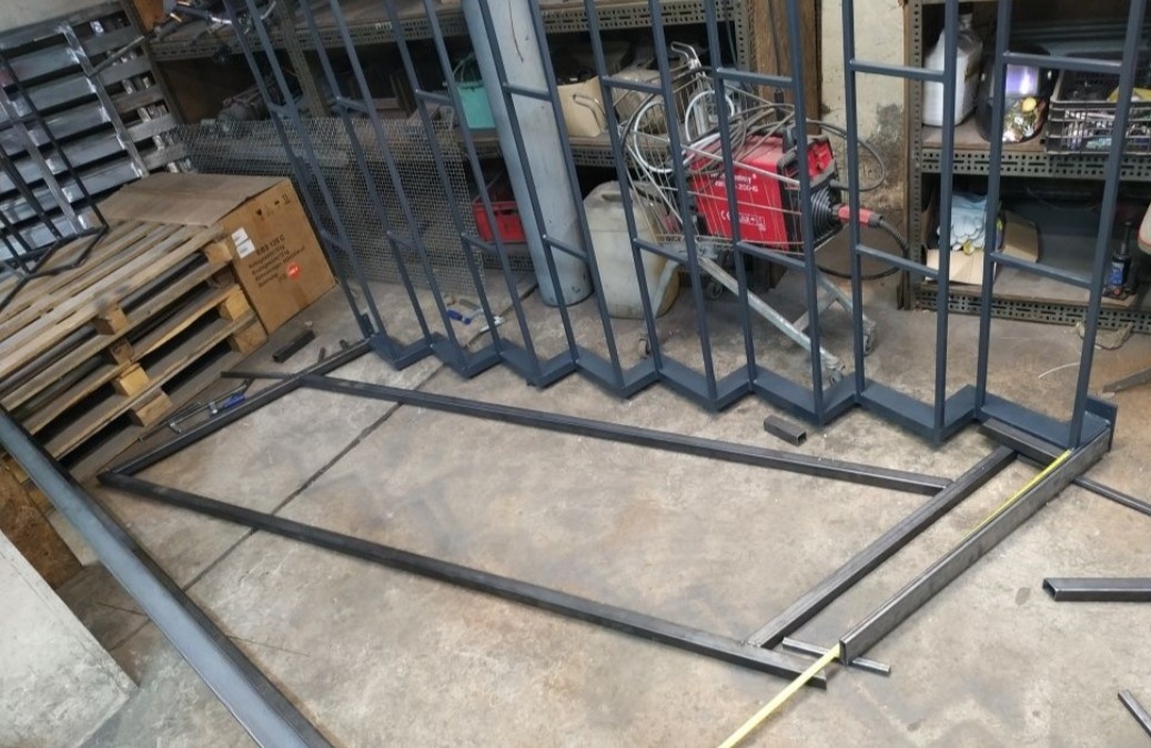 Konstrukcja metalowa w warsztacie: rama z profili kwadratowych i pionowe elementy z motywem schodkowym, spawarka w tle, widoczna taśma miernicza.