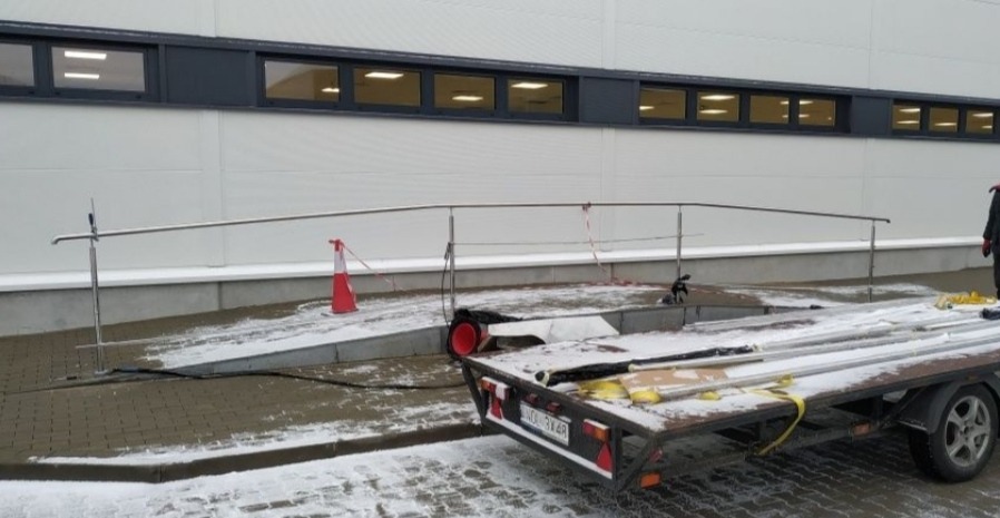 Montaż tymczasowej balustrady nierdzewnej na rampie, widoczna przyczepka z elementami konstrukcyjnymi, pomarańczowy pachołek, śnieg na podłożu.
