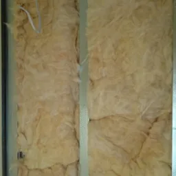 Poddasze w trakcie ocieplania wełną mineralną, widoczne profile metalowe i przewody elektryczne, ciepły odcień wełny.