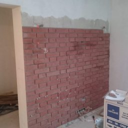 Wojsław - Ściana w trakcie wykańczania z cegieł dekoracyjnych w odcieniach czerwieni, widoczne przewody elektryczne, pozostałości kleju montażowego na górze ściany, wiadro z narzędziami i karton z ceramiką...
