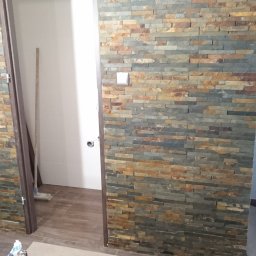 Wojsław - Ściana wykończona kamieniem dekoracyjnym w łazience, widoczne narzędzia glazurnicze i worek z zaprawą, wnętrze w trakcie prac wykończeniowych.