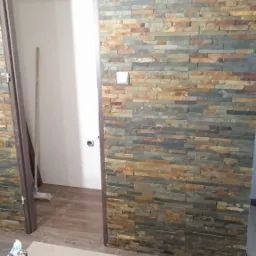 Ściana wykończona kamieniem dekoracyjnym w łazience, widoczne narzędzia glazurnicze i worek z zaprawą, wnętrze w trakcie prac wykończeniowych.