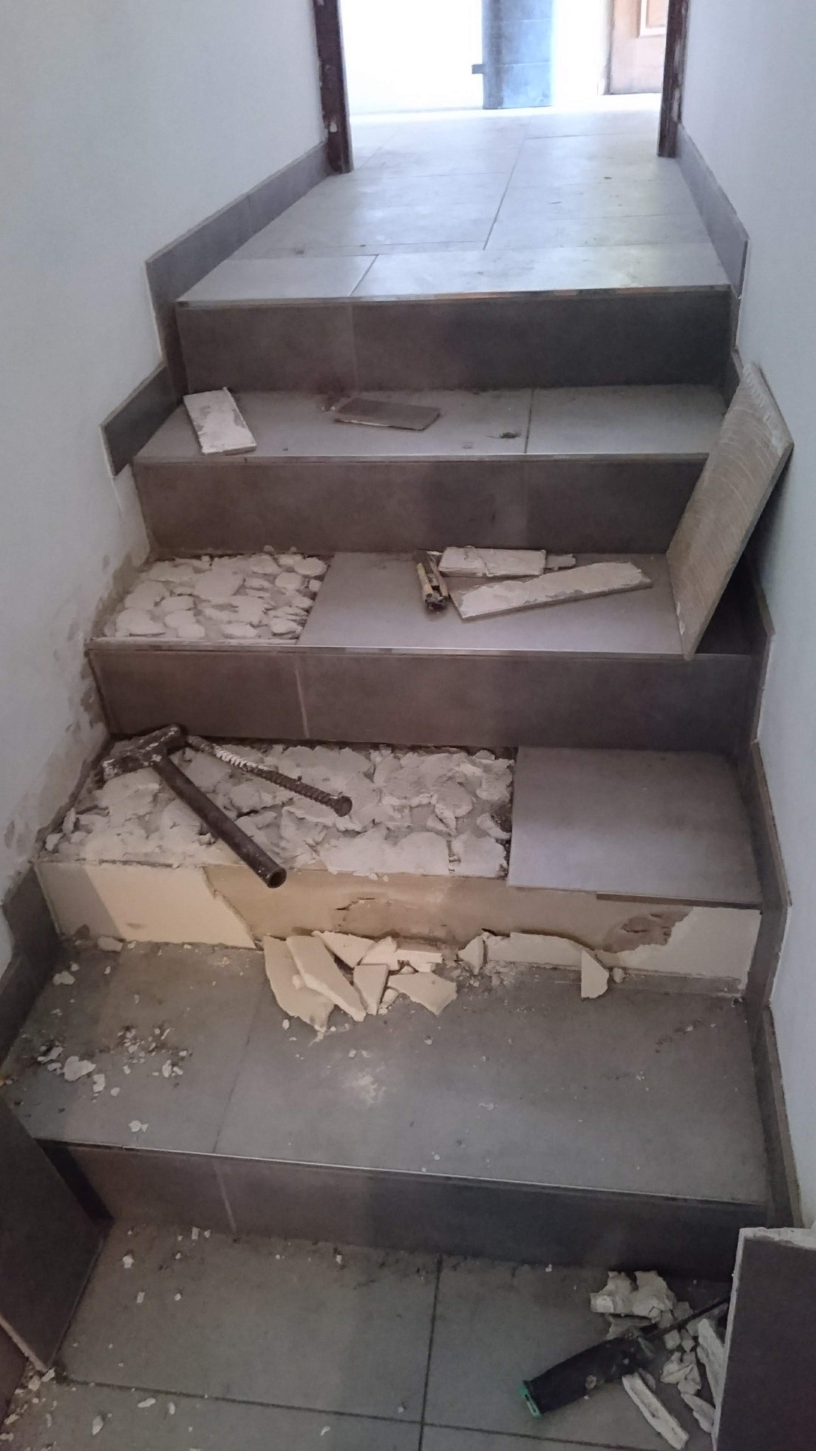 Schody w trakcie remontu, z częściowo usuniętymi płytkami, widoczne narzędzia glazurnicze i gruz.