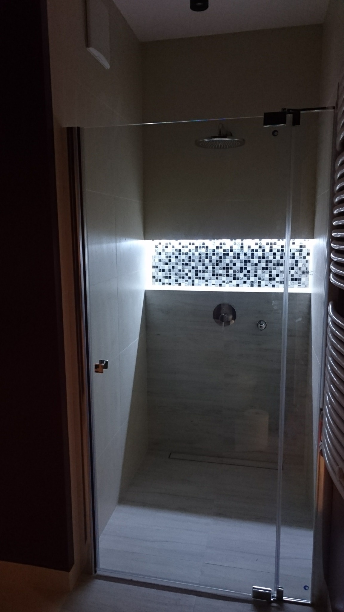 Nowoczesna kabina prysznicowa z deszczownicą i dekoracyjną mozaiką podświetloną LED, widok z zewnątrz przez szklane drzwi.
