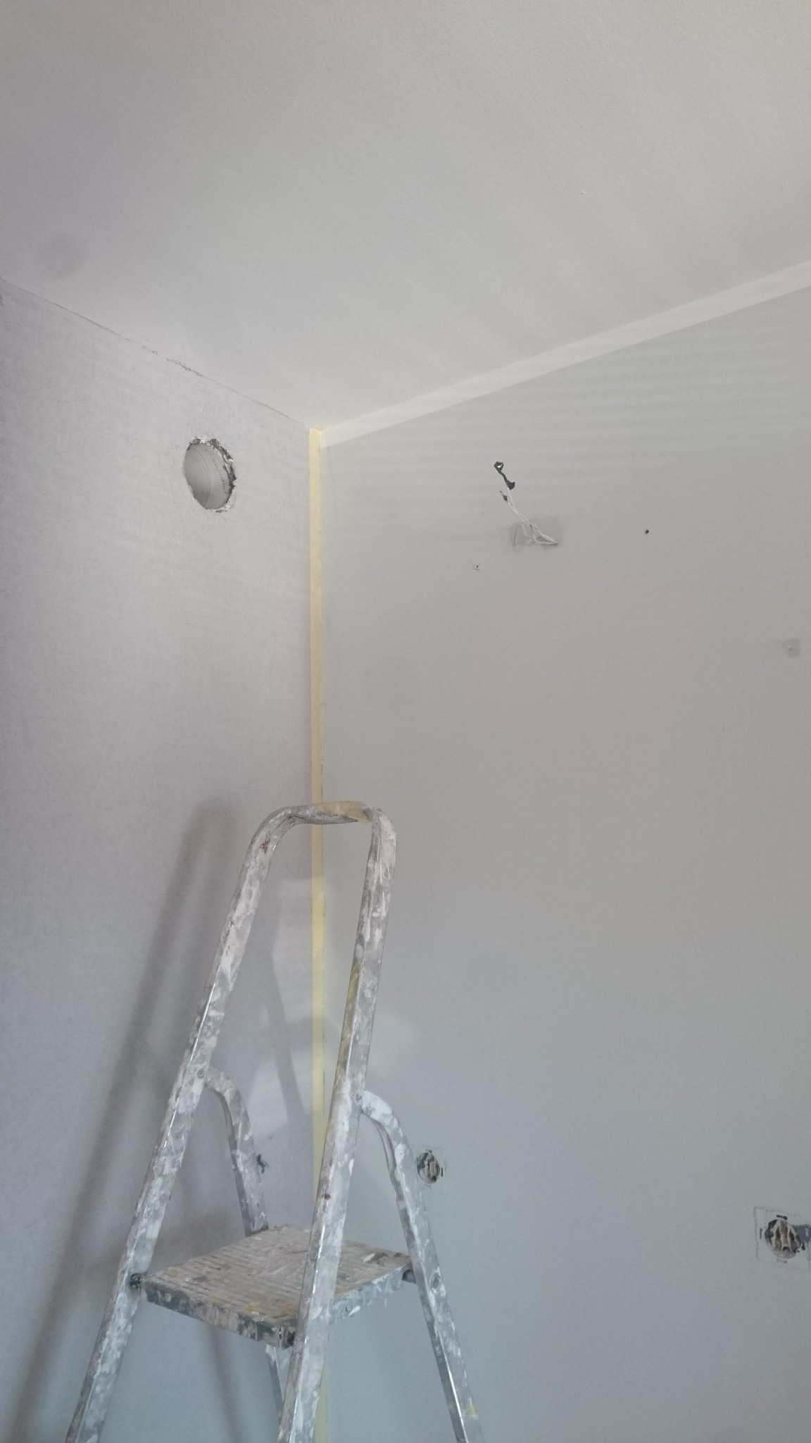 Świeżo pomalowane, jasnoszare wnętrze z widoczną drabiną malarską, zabezpieczeniami taśmą malarską przy suficie i wokół otworu wentylacyjnego, oraz niezamaskowanymi gniazdkami elektrycznymi...