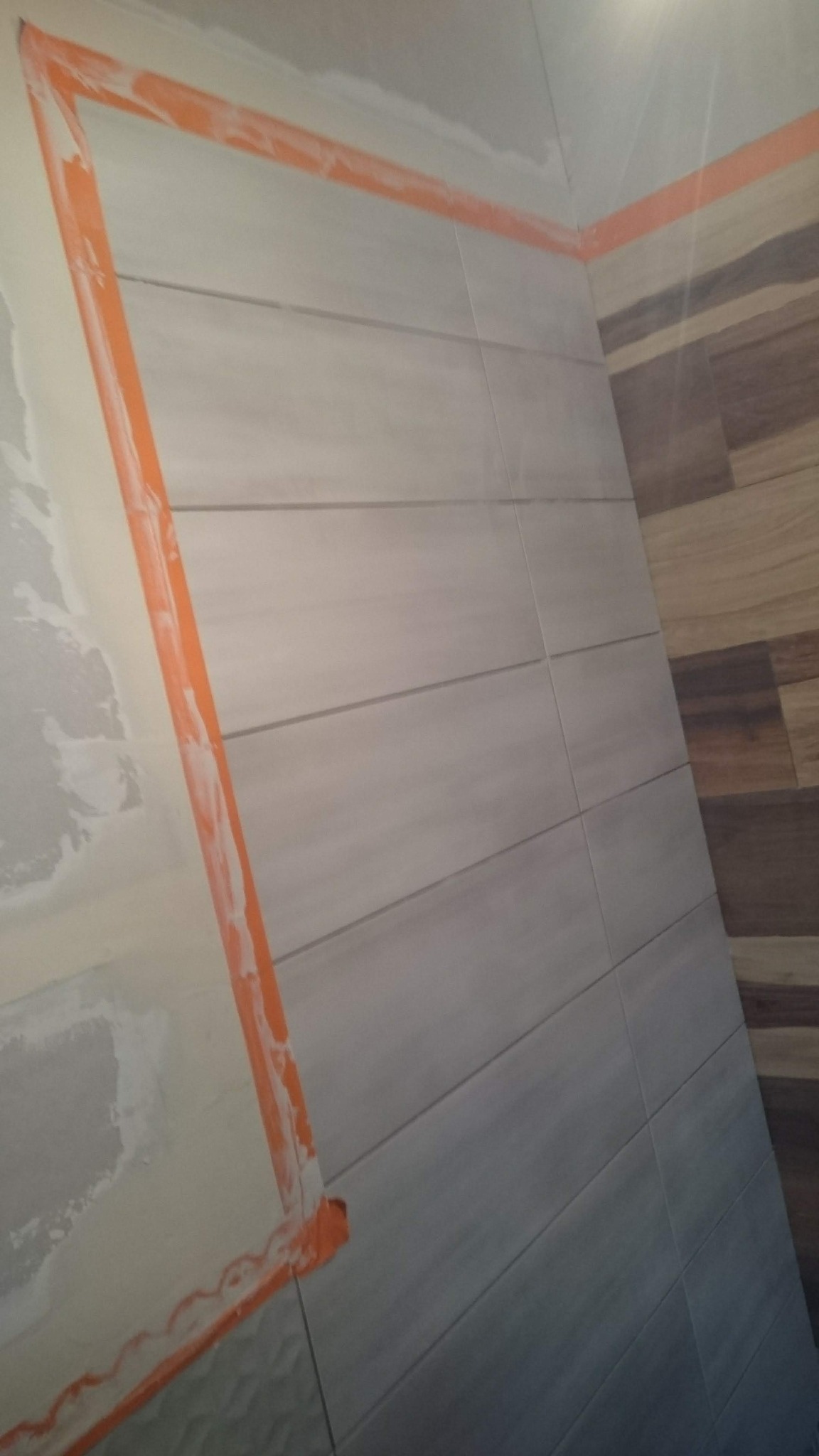 Płytki ceramiczne imitujące drewno ułożone na ścianie w łazience, zabezpieczone pomarańczową taśmą malarską przed dalszymi pracami wykończeniowymi, widoczne ślady zaprawy na ścianie obok.