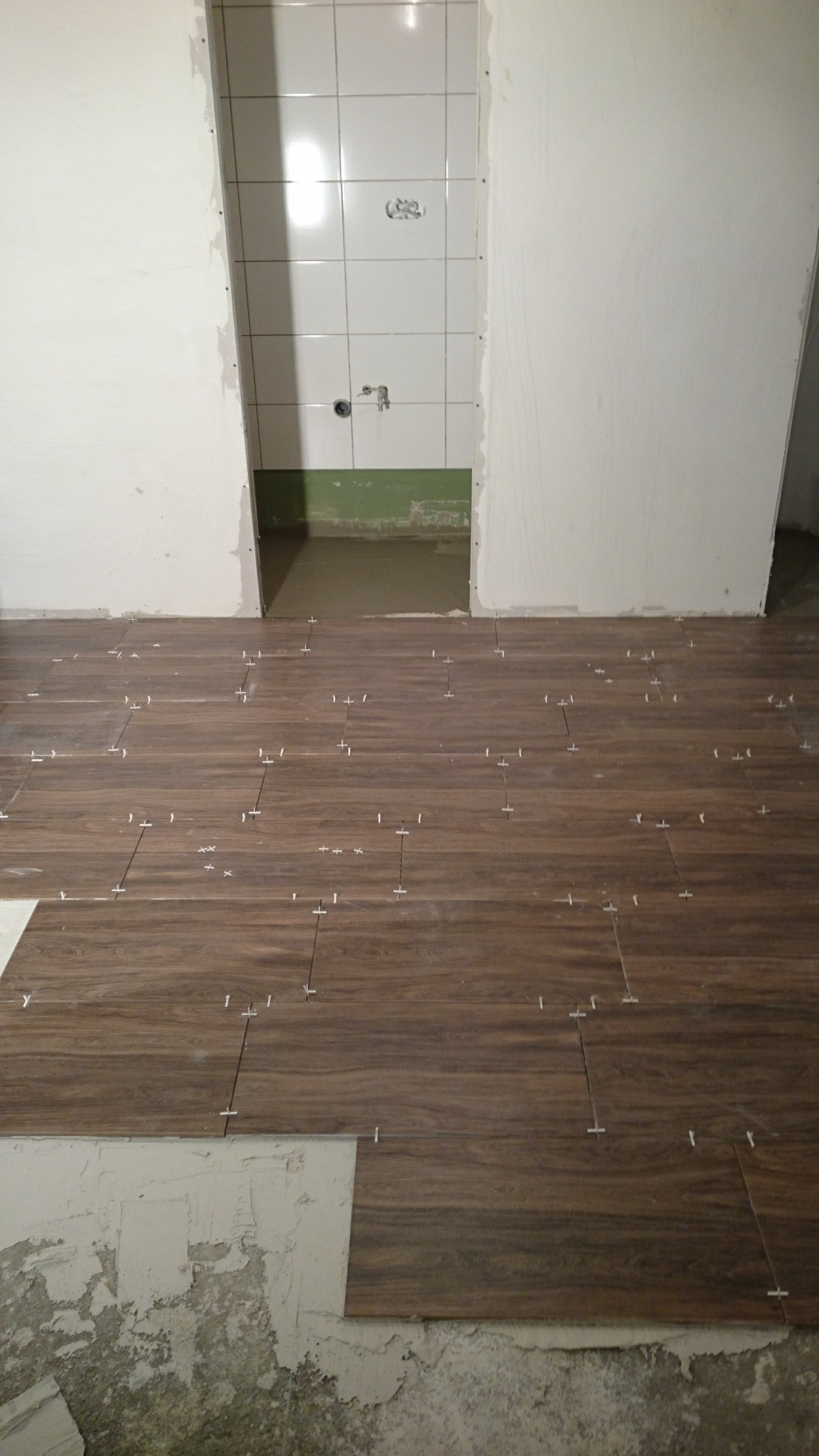 Proces układania brązowych płytek podłogowych imitujących drewno w łazience, z widocznymi krzyżykami dystansowymi i fragmentami ścian przygotowanych do dalszych prac wykończeniowych.
