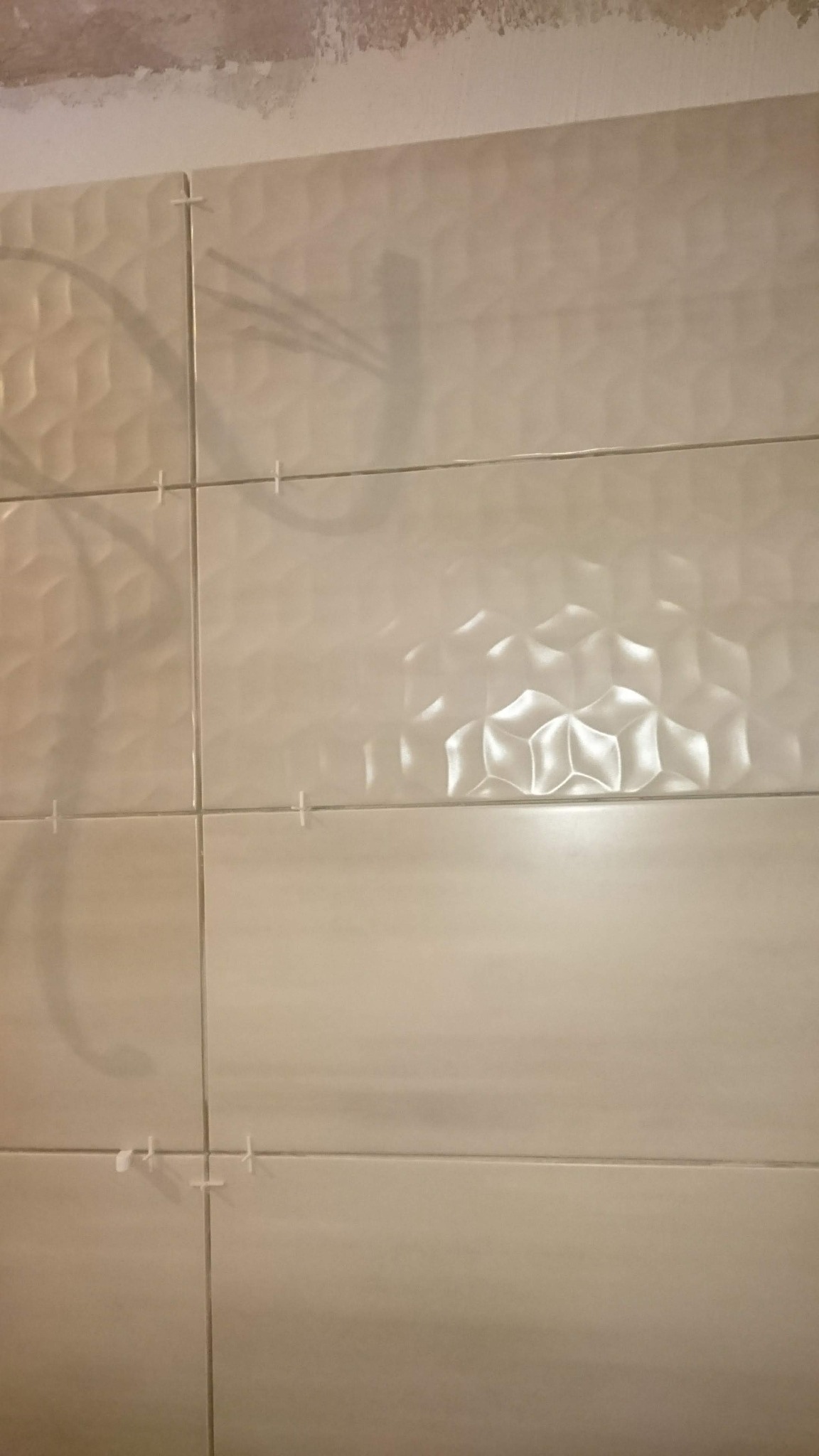 Świeżo ułożone, beżowe płytki ceramiczne z geometrycznym wzorem 3D na ścianie, z widocznymi krzyżykami dystansowymi i fragmentem surowego wykończenia sufitu.