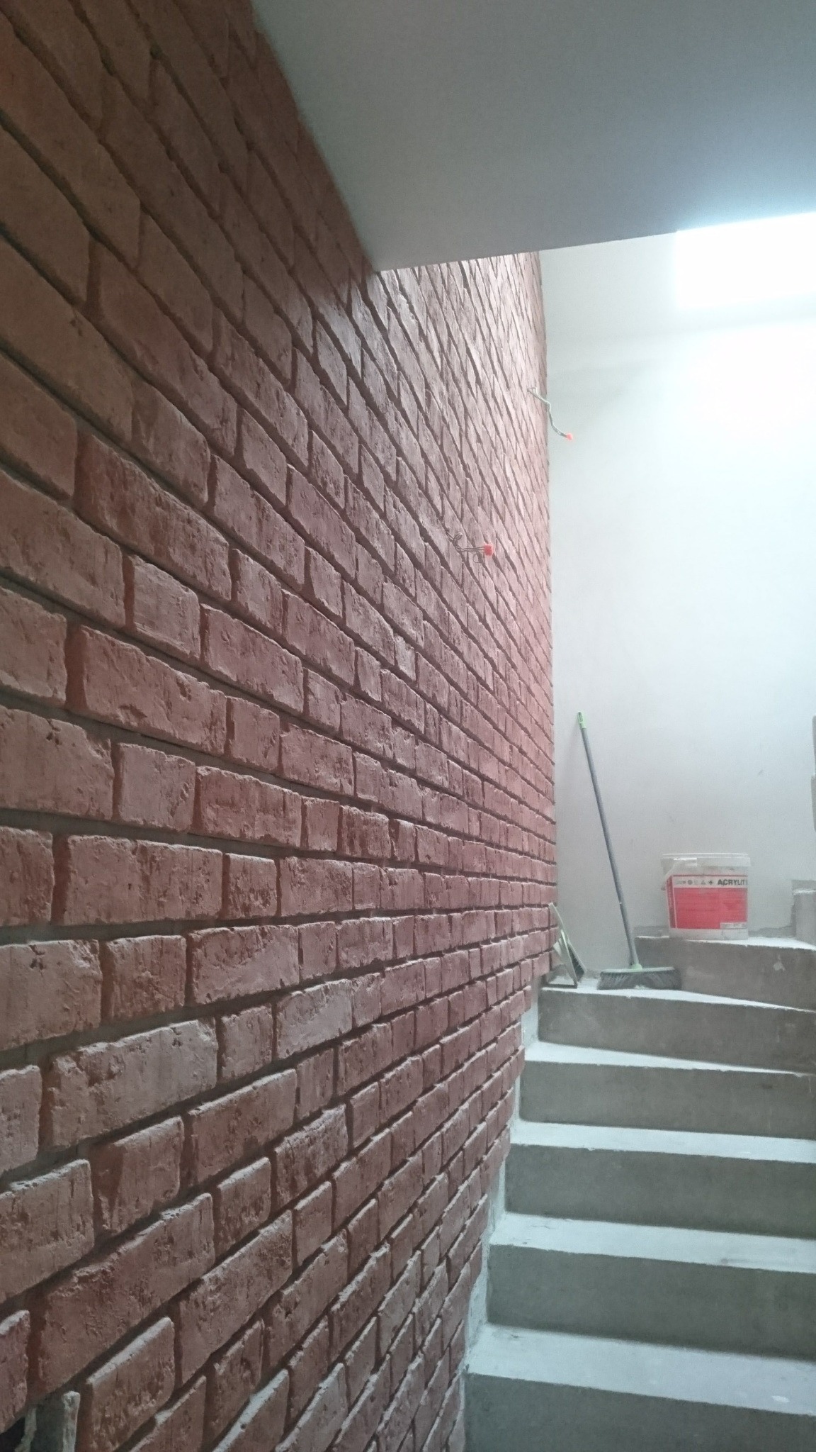 Ściana z cegły klinkierowej na klatce schodowej, widoczna puszka z farbą i zmiotka. Imitacja cegły na ścianie wewnętrznej.