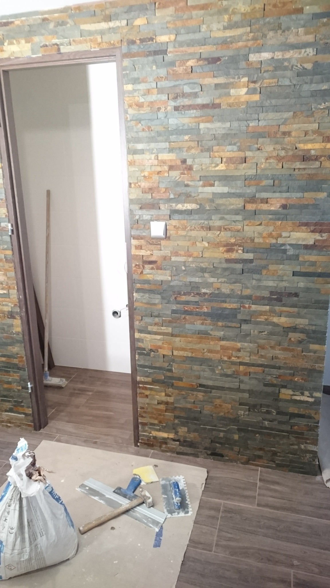 Ściana wykończona kamieniem dekoracyjnym w łazience, widoczne narzędzia glazurnicze i worek z zaprawą, wnętrze w trakcie prac wykończeniowych.