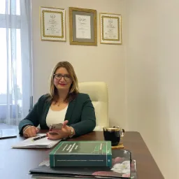 Kobieta w okularach i zielonej marynarce siedzi za biurkiem z dokumentami, smartfonem, książką o własności lokali i filiżanką; na ścianie wiszą trzy oprawione licencje zarządcy nieruchomości.