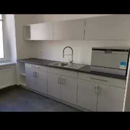 Szare szafki kuchenne z ciemnym blatem, chromowanym kranem i zintegrowaną zmywarką w nowym mieszkaniu, widok z przodu.