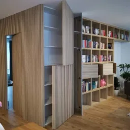 Nowoczesna zabudowa z drewnianymi lamelami, ukrywająca drzwi i szafę, połączona z regałem na książki w salonie z drewnianą podłogą.