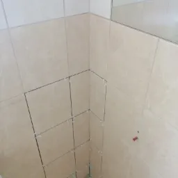 Beżowe płytki ścienne w trakcie układania w rogu pomieszczenia, widoczne krzyżyki dystansowe i poziomowanie, częściowo odbicie w lustrze.