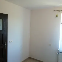 Pomalowane na biało ściany w rogu pokoju z ciemnymi drzwiami po lewej, widoczne gniazdka elektryczne, grzejnik pod oknem i ceramiczna podłoga w odcieniach szarości.