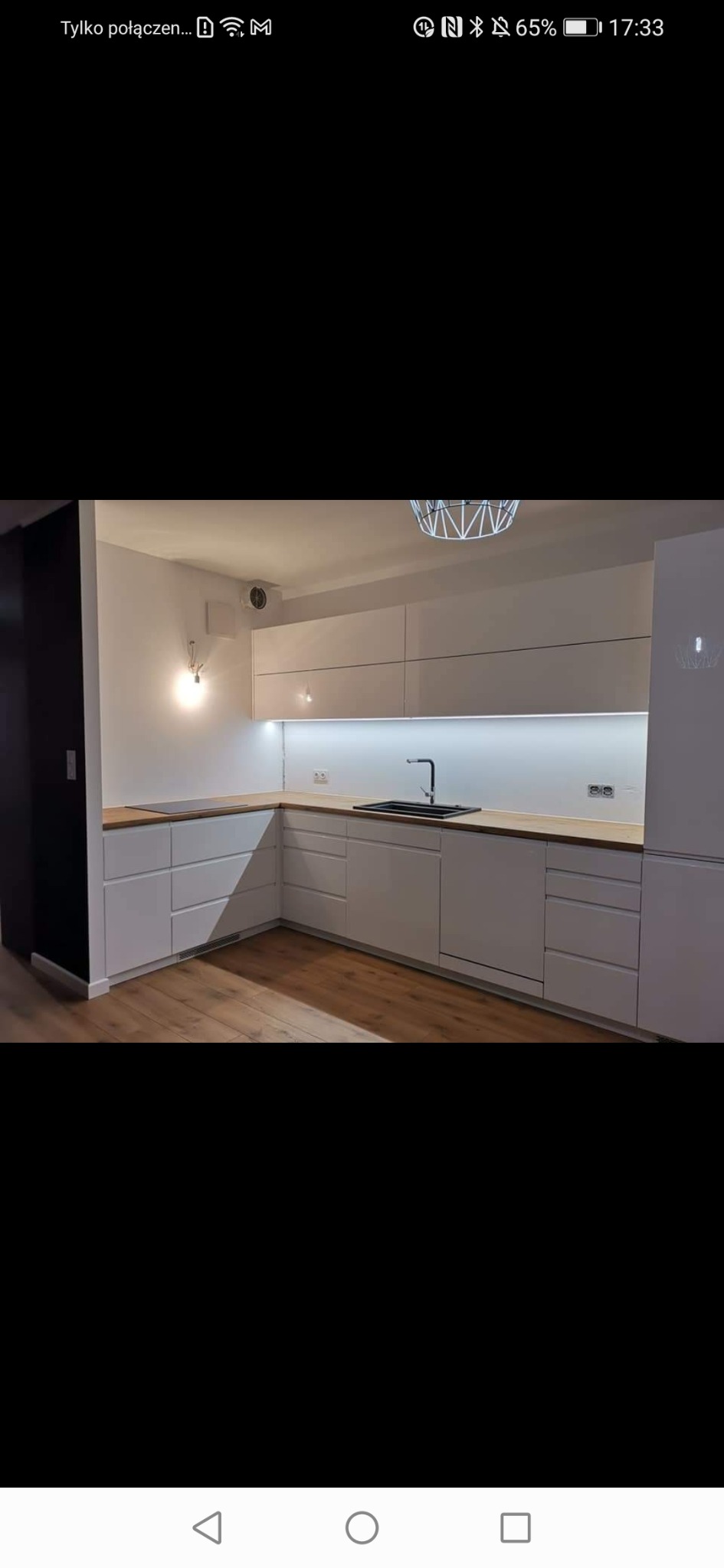 Nowoczesna kuchnia na wymiar z białymi frontami, drewnianym blatem i czarną armaturą, oświetlona listwą LED pod szafkami wiszącymi.