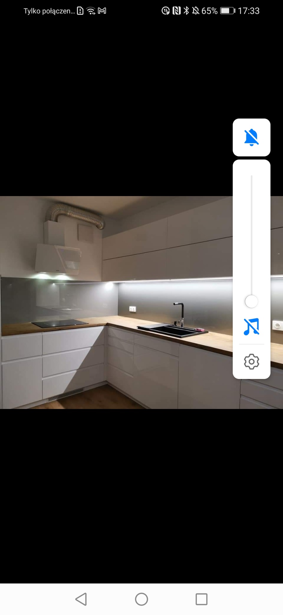 Nowoczesna, biała kuchnia z drewnianym blatem, czarnym zlewem i płytą indukcyjną. Oświetlenie LED pod szafkami górnymi i minimalistyczny okap.