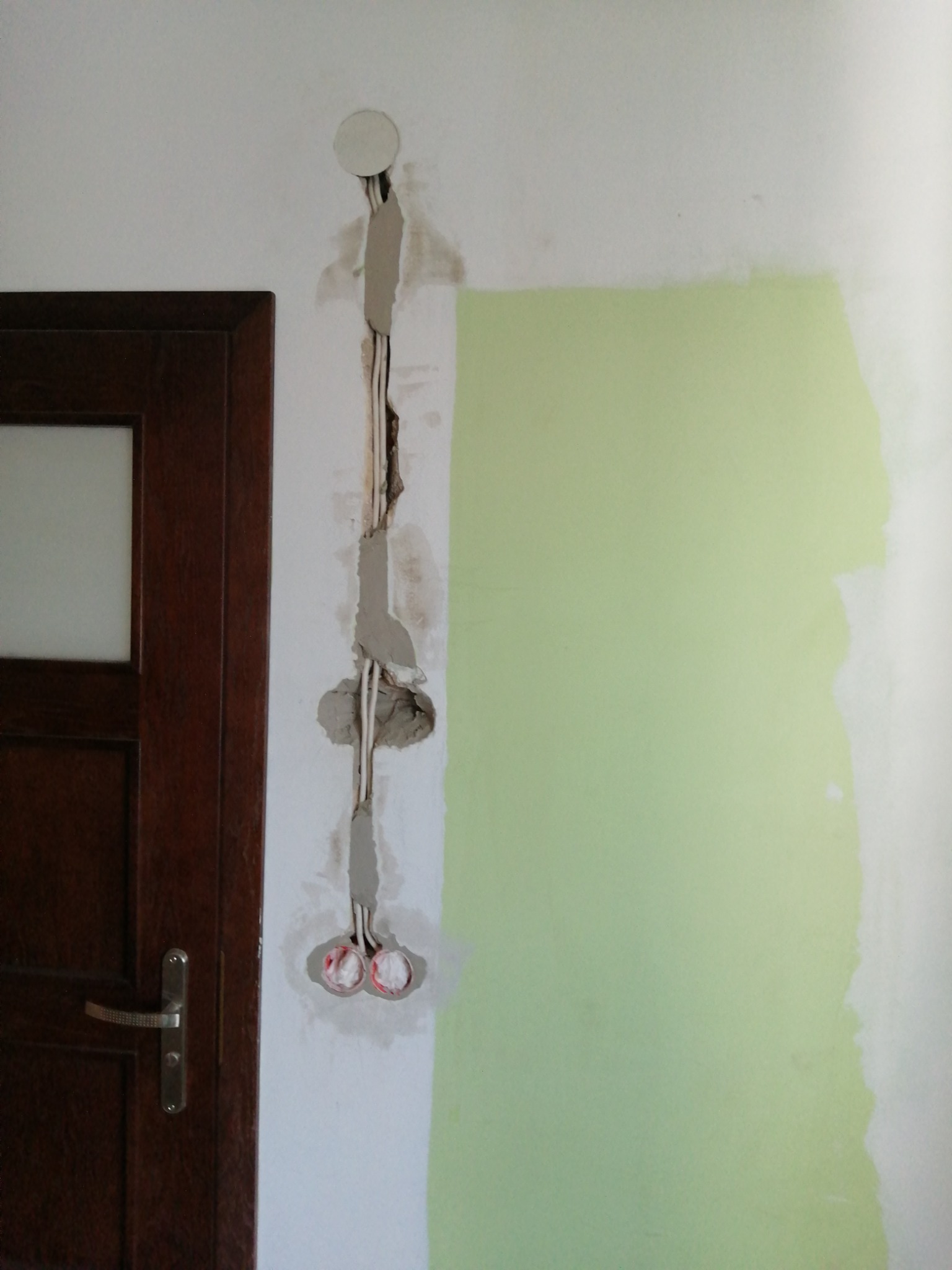 Fragment ściany z uszkodzonym tynkiem, widoczne przewody elektryczne, częściowo pomalowana na jasnozielony kolor, obok ciemne drzwi z klamką.