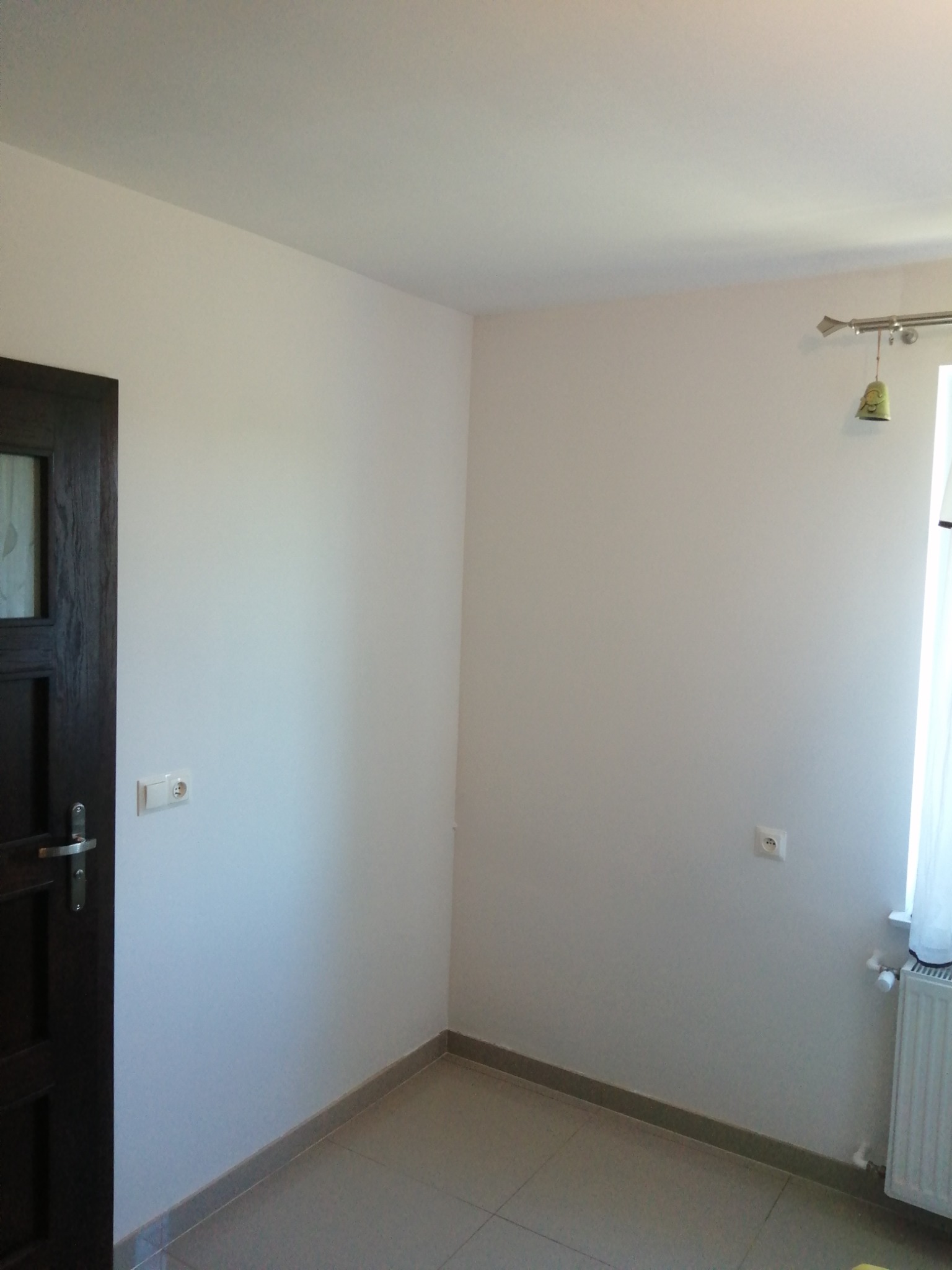 Pomalowane na biało ściany w rogu pokoju z ciemnymi drzwiami po lewej, widoczne gniazdka elektryczne, grzejnik pod oknem i ceramiczna podłoga w odcieniach szarości.