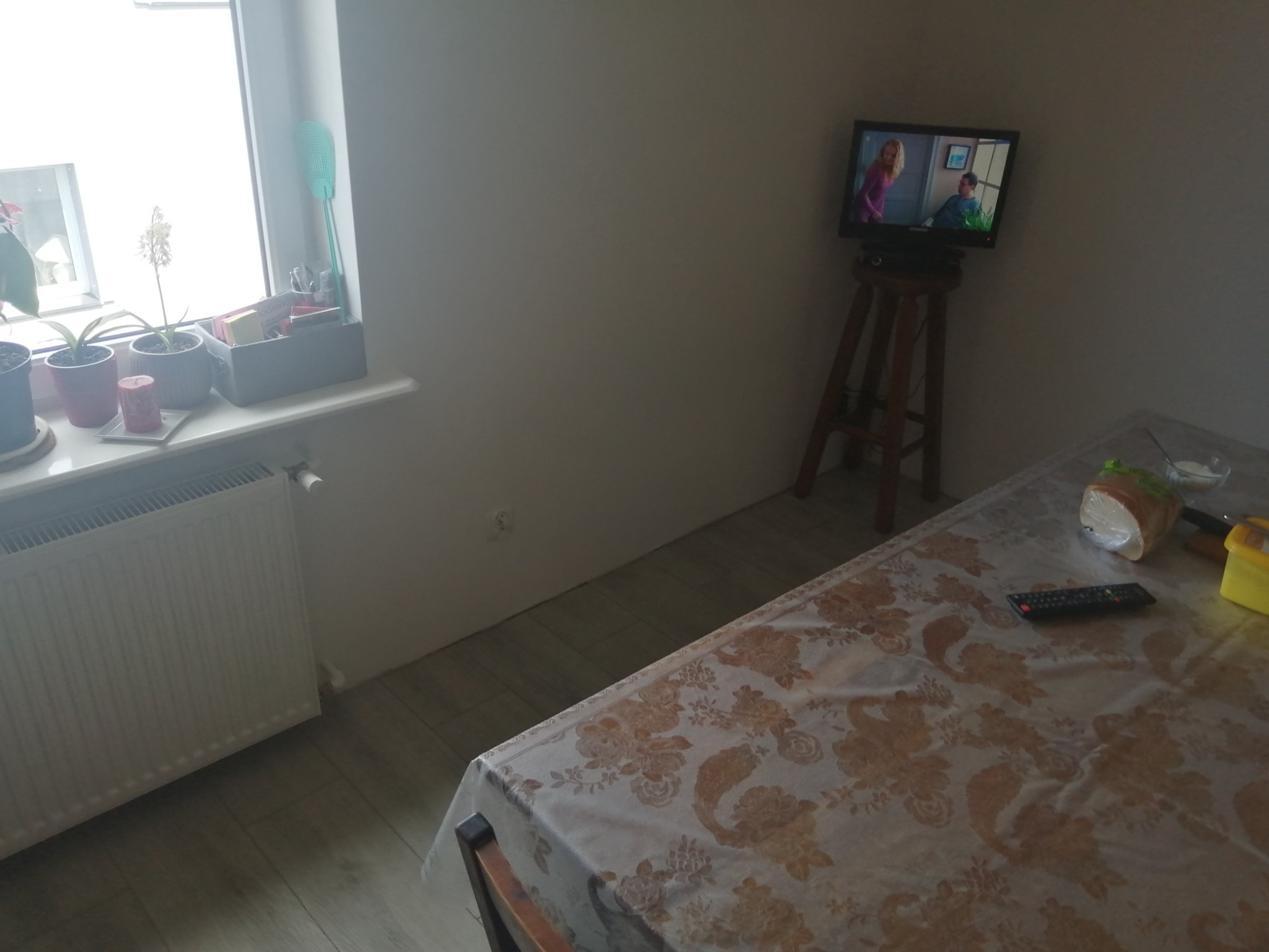 Pokój z telewizorem na drewnianym stołku, grzejnikiem pod oknem i stołem nakrytym obrusem w kwiaty