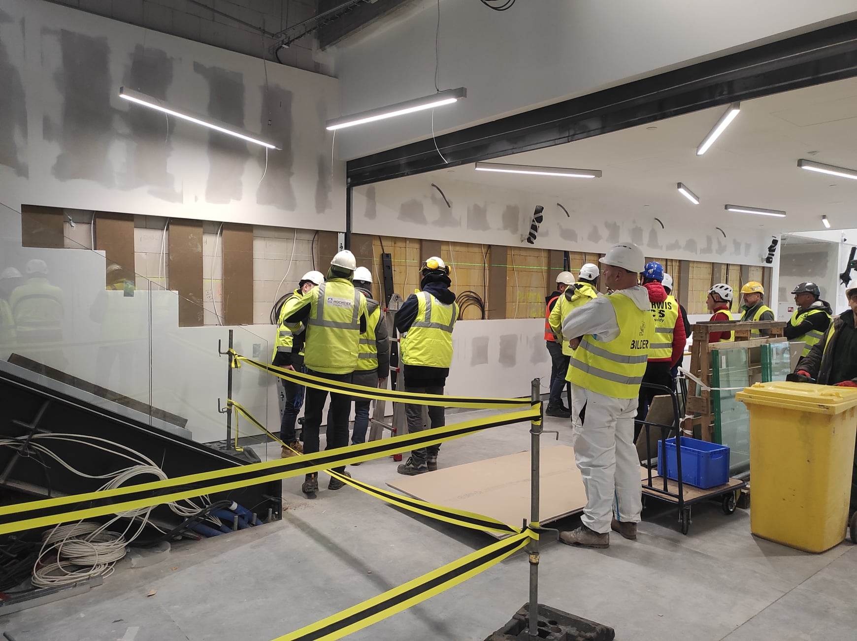Wnętrze w trakcie wykańczania: grupa pracowników budowlanych w kaskach i odblaskowych kamizelkach omawia montaż płyt kartonowo-gipsowych na ścianie, widoczne elementy instalacji elektrycznej...