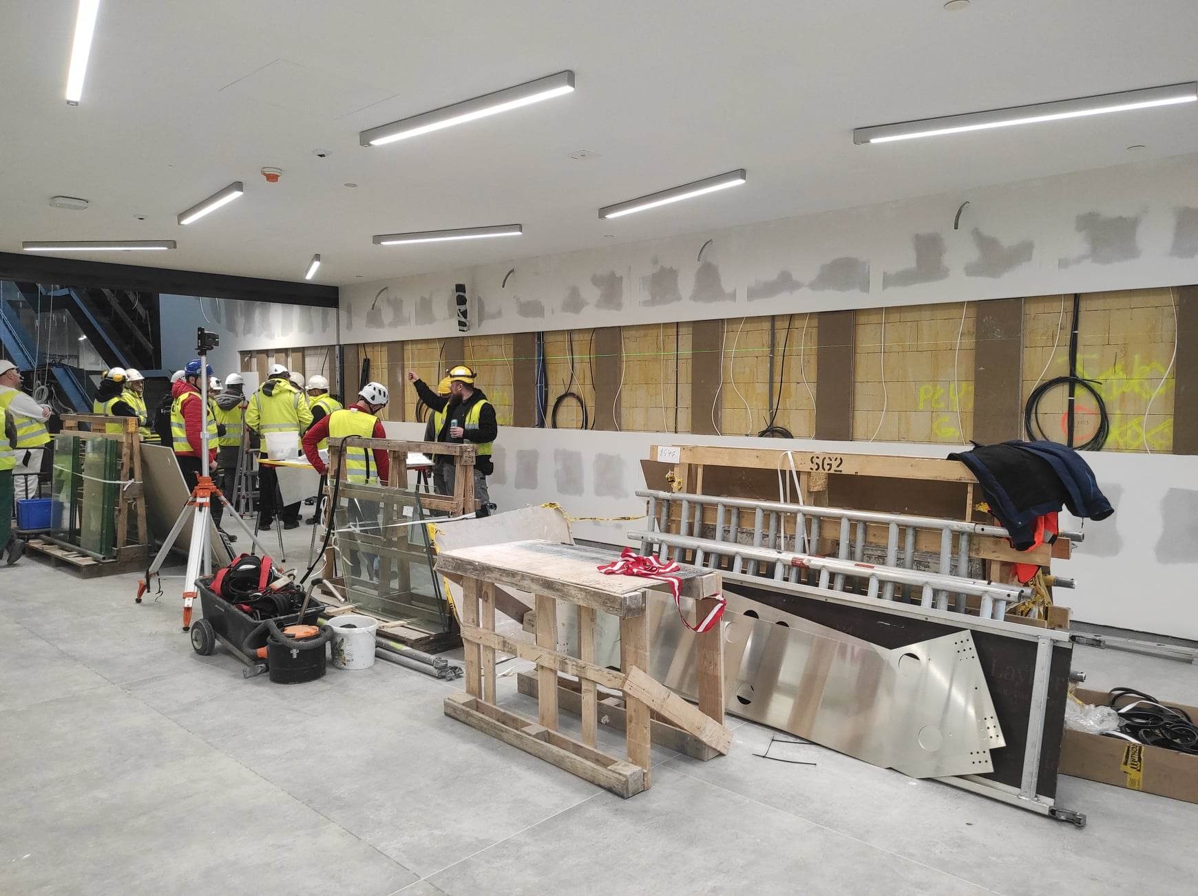 Wnętrze biura w trakcie remontu, widoczni pracownicy w kaskach i kamizelkach odblaskowych, przygotowujący się do montażu ścian działowych, z widocznymi elementami konstrukcyjnymi, przewodami...