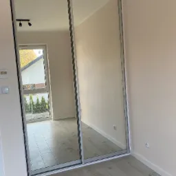 Szafa wnękowa z przesuwnymi drzwiami lustrzanymi, widoczna na tle jasnych ścian i drewnianej podłogi, z odbiciem okna i lamp sufitowych.