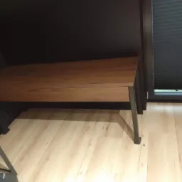 Drewniany blat biurka na metalowych nogach, zamontowany w rogu pokoju z ciemną ścianą i jasną podłogą z paneli, widoczne rolety w oknie.