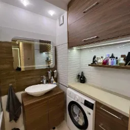 Nowoczesna łazienka z drewnianymi szafkami, umywalką nablatową, podświetlanym lustrem LED, pralką Electrolux i półką z kosmetykami.