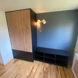 Wysoka szafa z drewnianymi frontami i czarnymi bokami stoi obok czarnej szafki z półkami, oświetlona przez lampę ścienną, na tle szarej ściany i drewnianej podłogi.