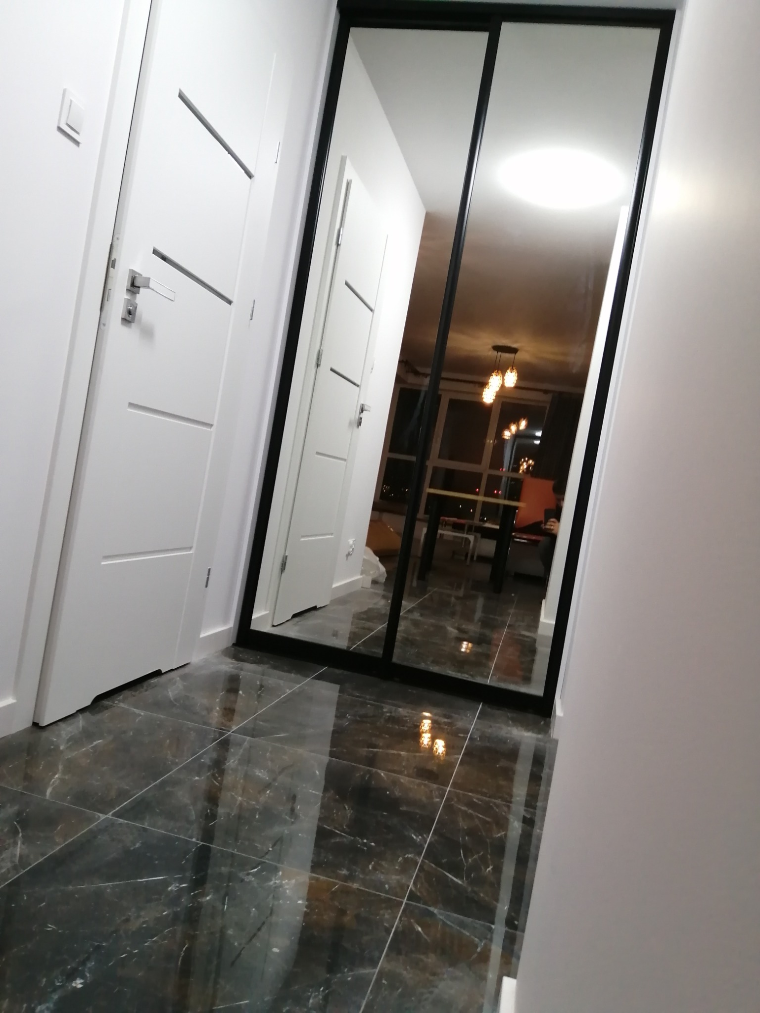Nowoczesna szafa wnękowa z przesuwnymi drzwiami lustrzanymi i czarną ramą w korytarzu z ciemną, marmurkową podłogą; w lustrze odbija się oświetlony pokój z widoczną lampą wiszącą i oknem.