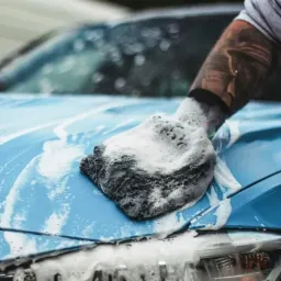 Błękitny samochód myty gąbką z dużą ilością piany, widoczna wytatuowana ręka osoby myjącej, w tle zamazane postacie.