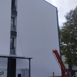 Ganz-bud - Świeżo pomalowana, jasna elewacja budynku wielorodzinnego z widocznym podnośnikiem koszowym, wiaderkami z farbą i wałkiem malarskim na trawniku.