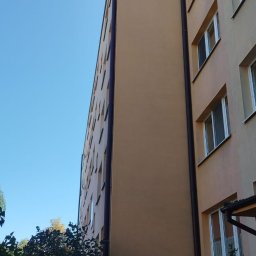 Ganz-bud - Wielopiętrowy budynek mieszkalny z pomarańczowo-beżową elewacją, widoczny z dołu pod kątem, z brązowymi rynnami i zielenią w tle.