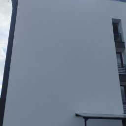 Ganz-bud - Fragment budynku wielorodzinnego z odnowioną, białą elewacją, widoczna drabina aluminiowa i pracownik w ciemnej koszulce przy podstawie ściany