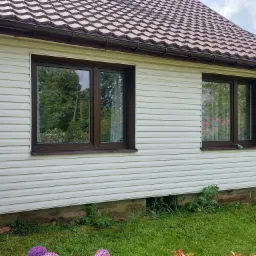 Dom z elewacją sidingową i brązowymi oknami, na parapecie leży płyn do szyb, a obok na trawie leży szczotka teleskopowa do mycia okien.