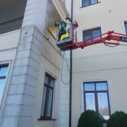 Pracownik na podnośniku koszowym czyści elewację budynku za pomocą myjki ciśnieniowej, strumień wody usuwa zabrudzenia z powierzchni.