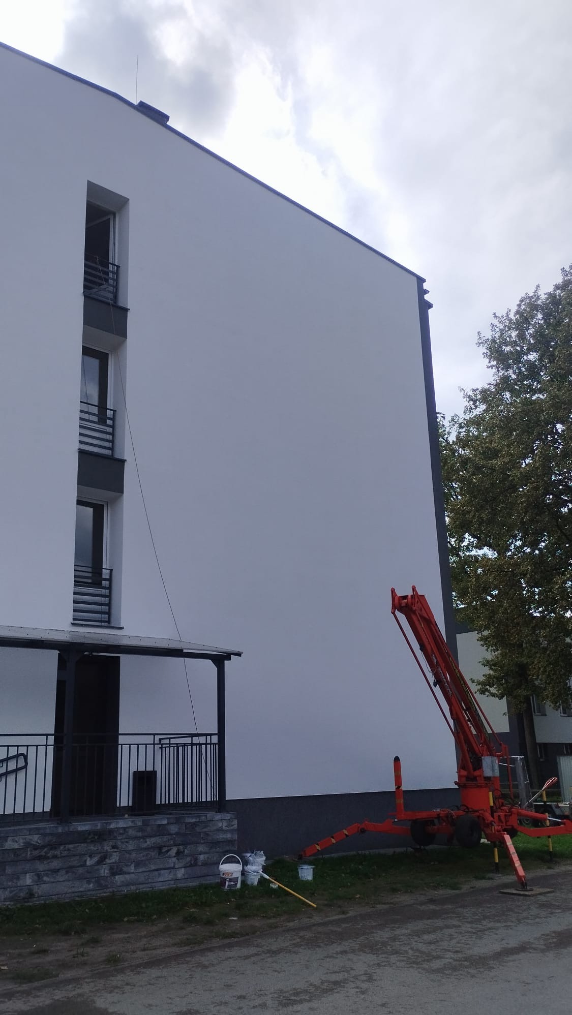 Świeżo pomalowana, jasna elewacja budynku wielorodzinnego z widocznym podnośnikiem koszowym, wiaderkami z farbą i wałkiem malarskim na trawniku.