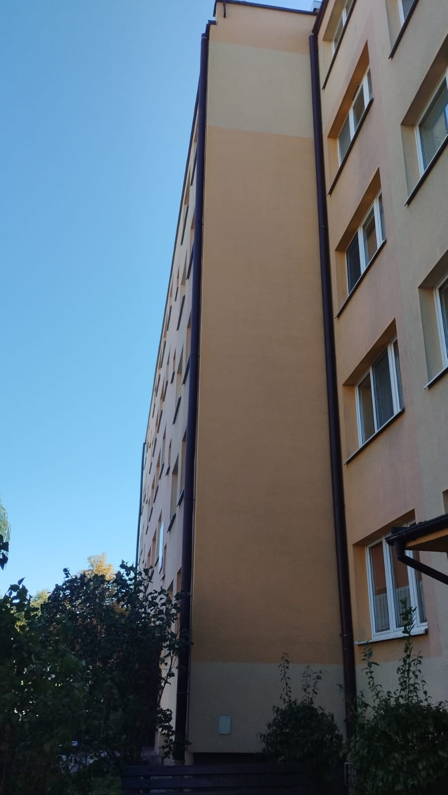 Wielopiętrowy budynek mieszkalny z pomarańczowo-beżową elewacją, widoczny z dołu pod kątem, z brązowymi rynnami i zielenią w tle.