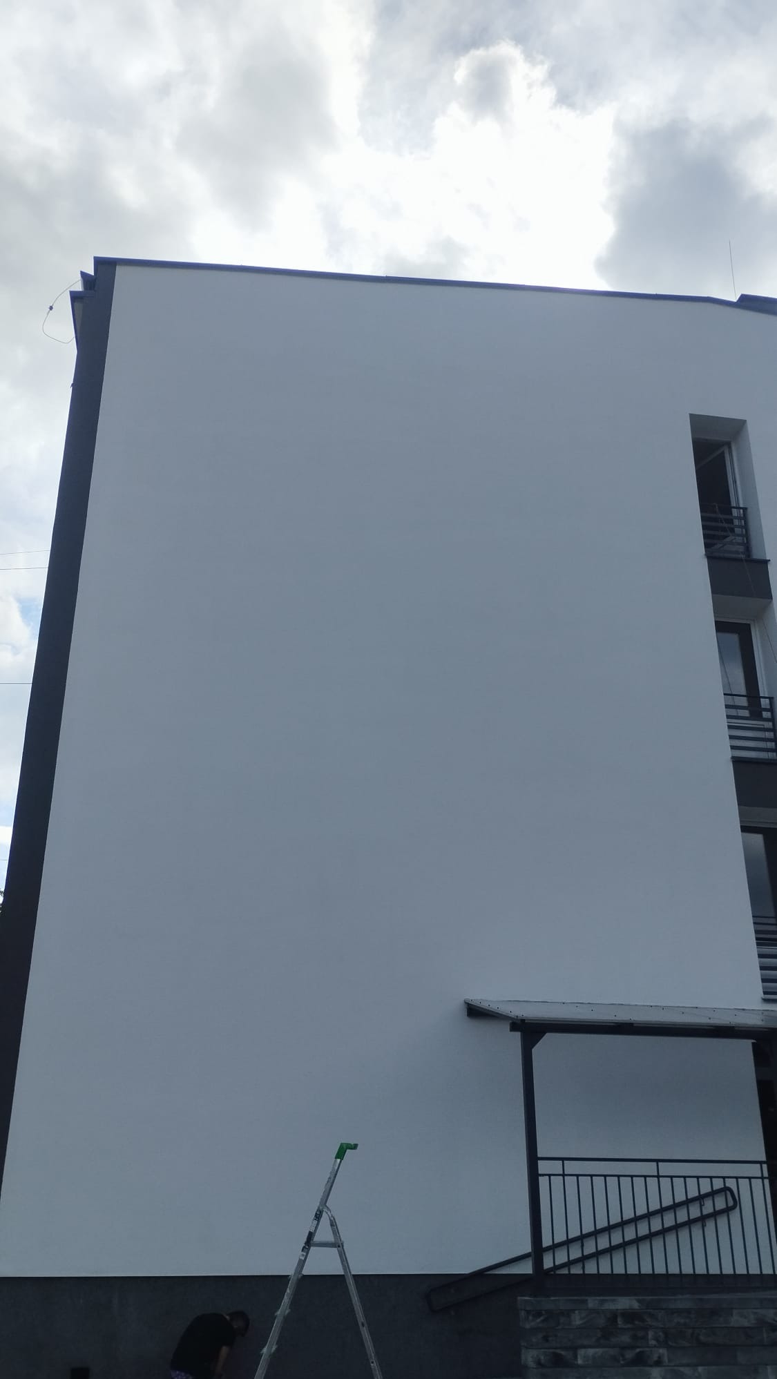 Fragment budynku wielorodzinnego z odnowioną, białą elewacją, widoczna drabina aluminiowa i pracownik w ciemnej koszulce przy podstawie ściany