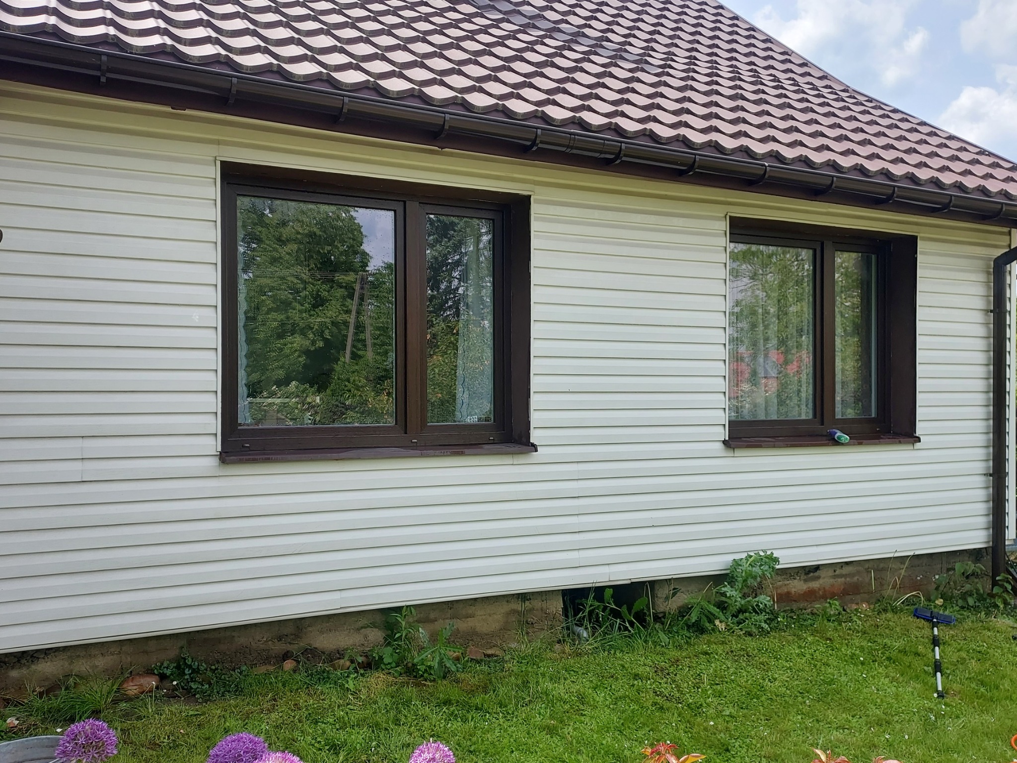 Dom z elewacją sidingową i brązowymi oknami, na parapecie leży płyn do szyb, a obok na trawie leży szczotka teleskopowa do mycia okien.