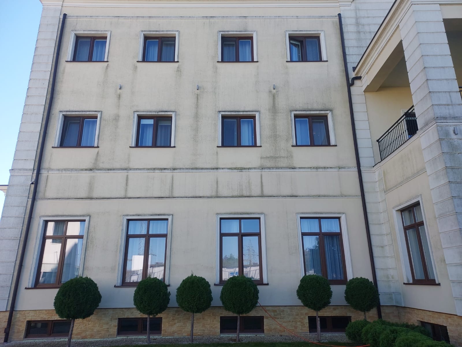 Elewacja budynku z widocznymi zabrudzeniami i plamami, brązowe ramy okienne, okrągłe krzewy ozdobne u podstawy, częściowo otwarte okno na wyższym piętrze.