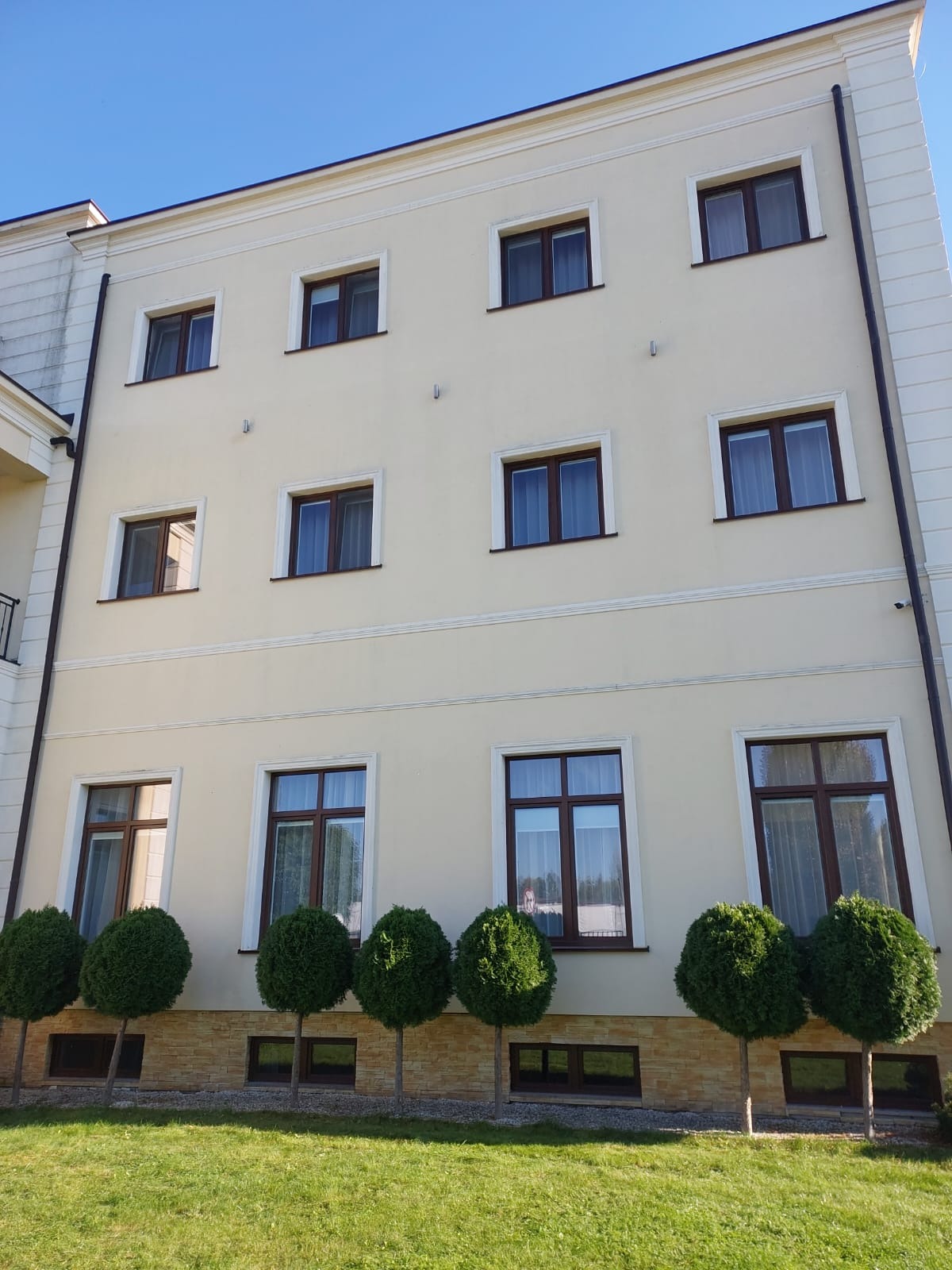 Elewacja budynku z kremową fasadą, brązowymi oknami i dekoracyjnymi elementami architektonicznymi, widok z poziomu trawnika z rzędem kulistych krzewów.
