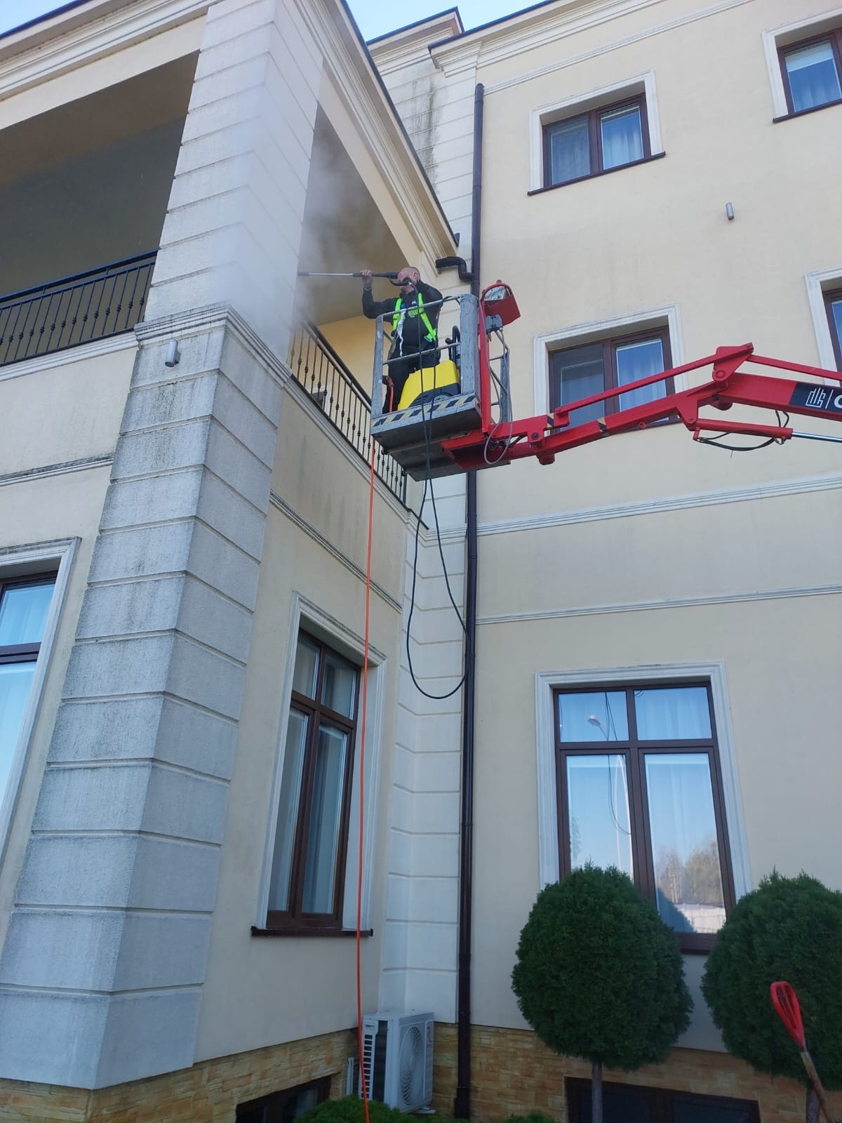 Pracownik na podnośniku koszowym czyści elewację budynku za pomocą myjki ciśnieniowej, strumień wody usuwa zabrudzenia z powierzchni.