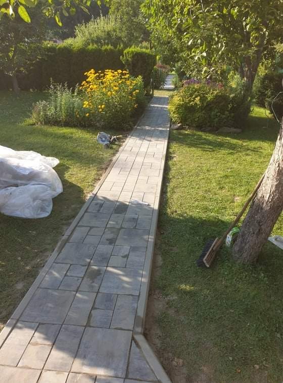 Ścieżka z szarej kostki brukowej prowadząca przez zielony ogród z kwitnącymi żółtymi i fioletowymi kwiatami, widoczny worek z materiałem budowlanym i szczotka oparta o drzewo.