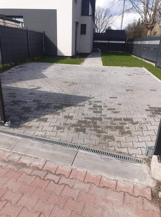 Ułożona szara kostka brukowa na podjeździe z odpływem liniowym, widoczny fragment czerwonej kostki przy krawędzi, w tle budynek z szarą elewacją i ogrodzenie.