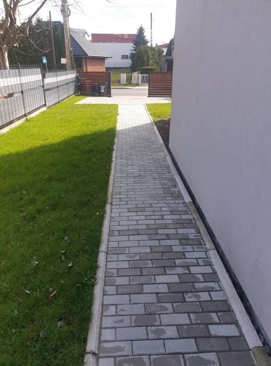 Ścieżka z szarej kostki brukowej prowadząca wzdłuż ściany budynku i trawnika, widok z góry.